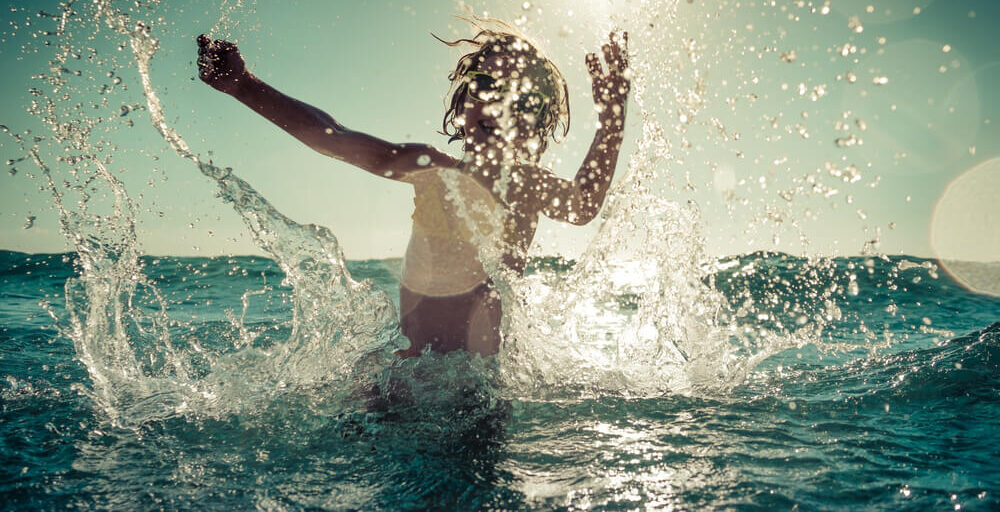 Dziecko w wodzie, na tle zachodzącego powoli słońca, rozchlapuje wokół krople wody