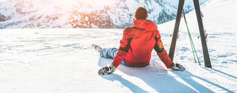 Mężczyzna w stroju narciarskim siedzący na stoku, patrzący na ośnieżone góry
